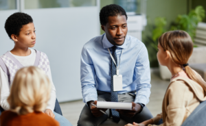 Man with a lanyard and clipboard leads a discussion with a small group of young students seated in a circle.