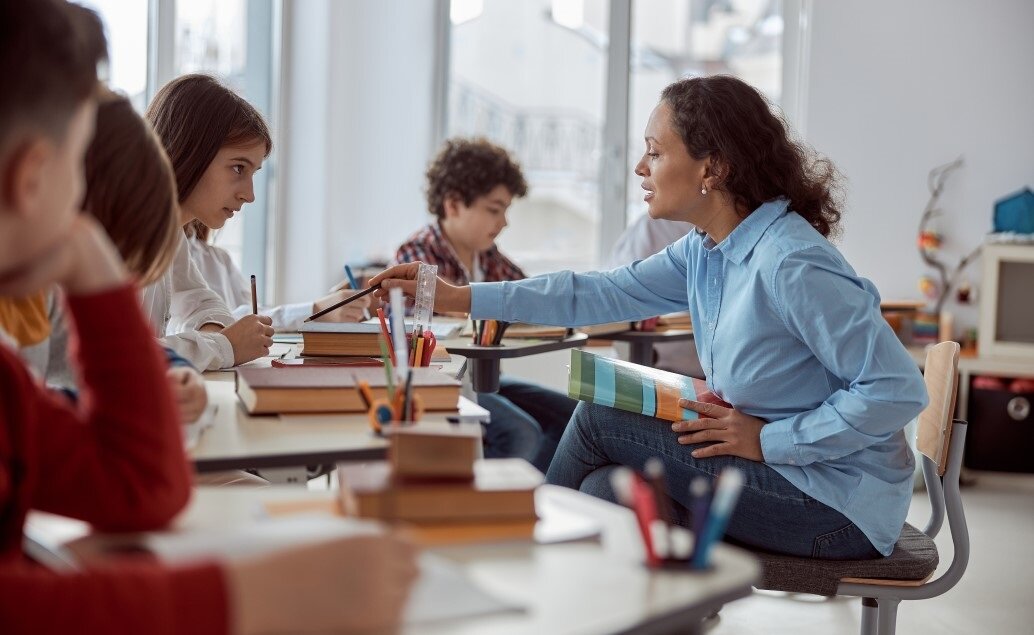 A possible new teacher is helping a student at a desk with other students at desks in the background.