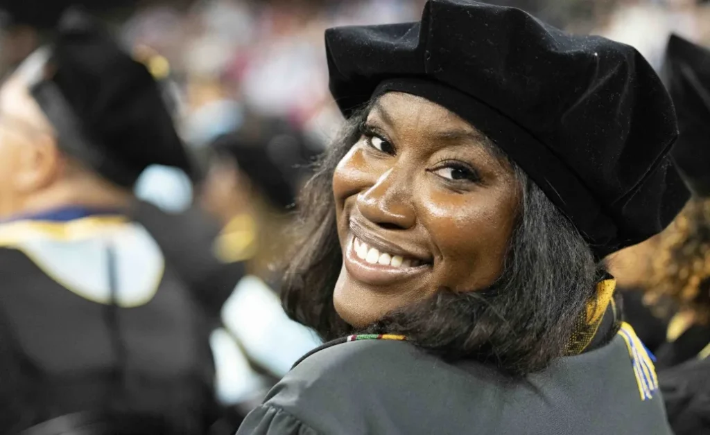 Graduate in a doctoral tam and gown looking back over her shoulder with a wide smile during a commencement ceremony.