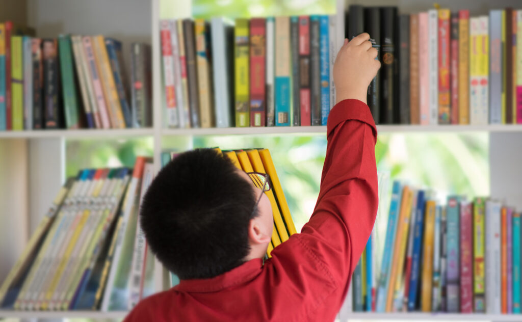 Student with glasses reaching up to a bookshelf filled with books in a library
