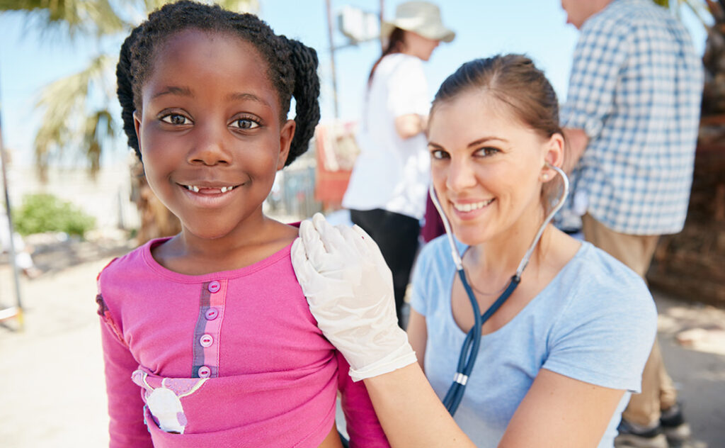 A young Black girl smiles at the camera while sitting on the lap of a white female, who is using a stethoscope to examine the girl at a public health community event