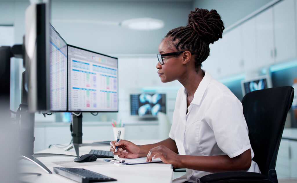 Black women works as a healthcare administration professional, taking notes from two computer monitors displaying data