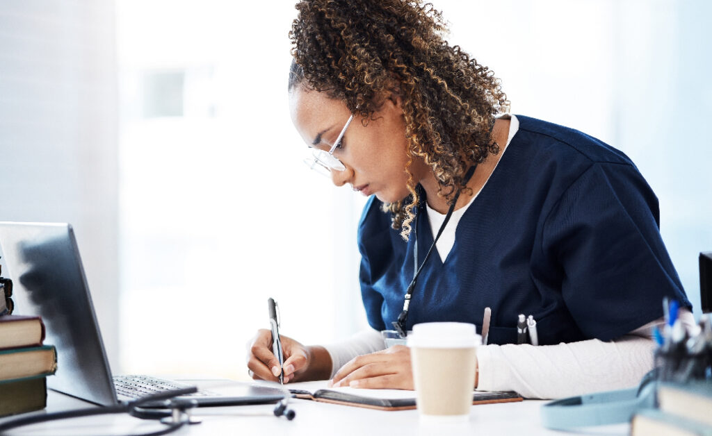 Young Black woman in scrubs writing in a notebook in front of a laptop