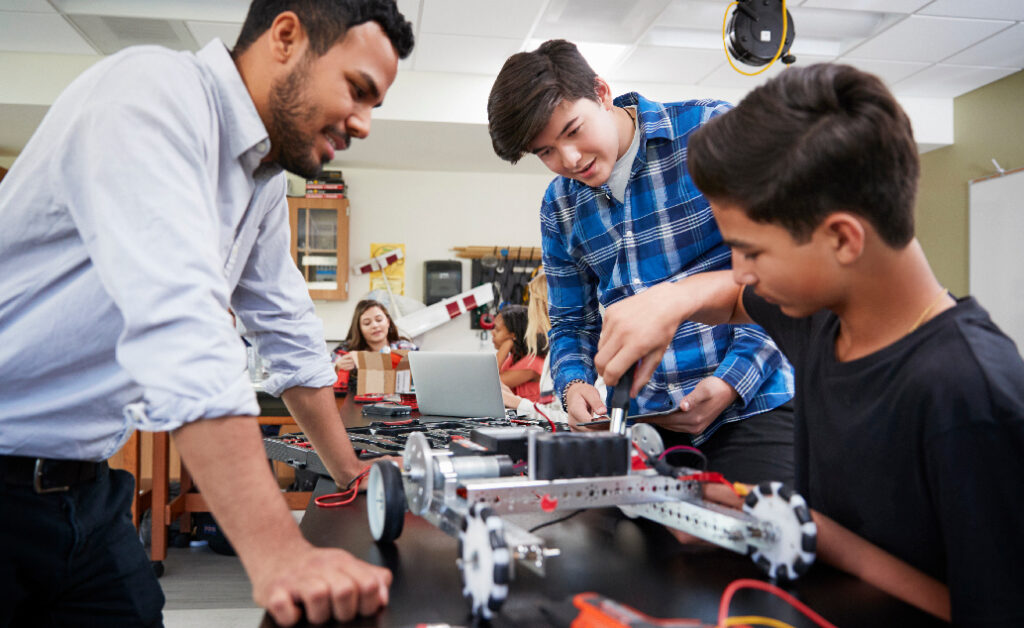 A male teacher oversees three students in a science classroom working on a robotics project