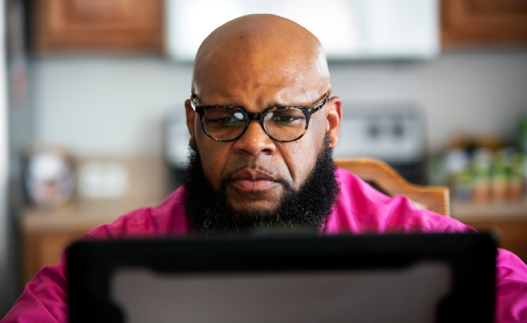 Man wearing glasses and a bright pink shirt focuses on his online education on laptop.