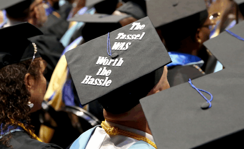 Photo of a row of graduates with a focus on a decorated grad cap that says, "The tassel was worth the hassle"