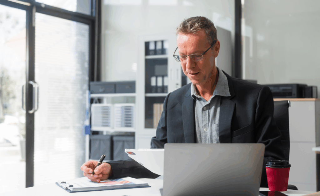 An older white male sits at a desk reviewing papers while sitting in front of a laptop