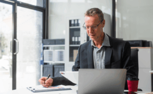 An older white male sits at a desk reviewing papers while sitting in front of a laptop