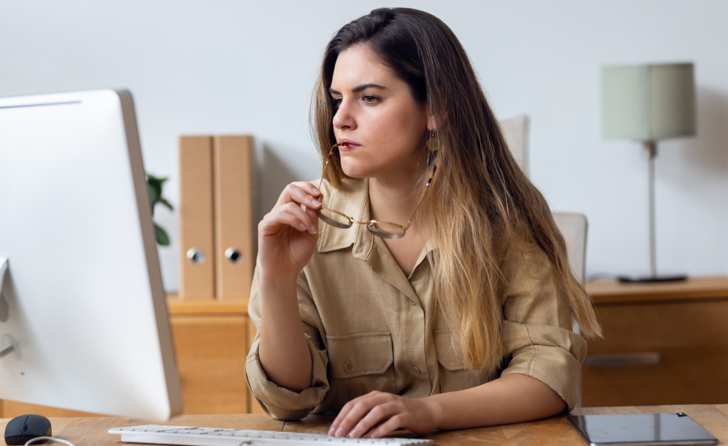 Woman working at a desk, looking thoughtfully at a computer screen.