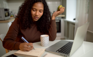 A woman taking notes on a notebook while studying on her laptop.