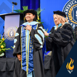 Two faculty members in academic regalia at a graduation ceremony, one being hooded on stage