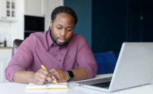 Man at a kitchen table taking notes in a notebook while studying online on his laptop.