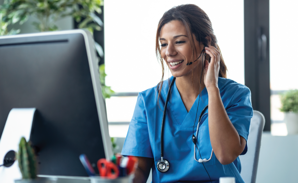 Smiling nurse in scrubs wearing a headset and stethoscope while working at a computer for a telehealth call.