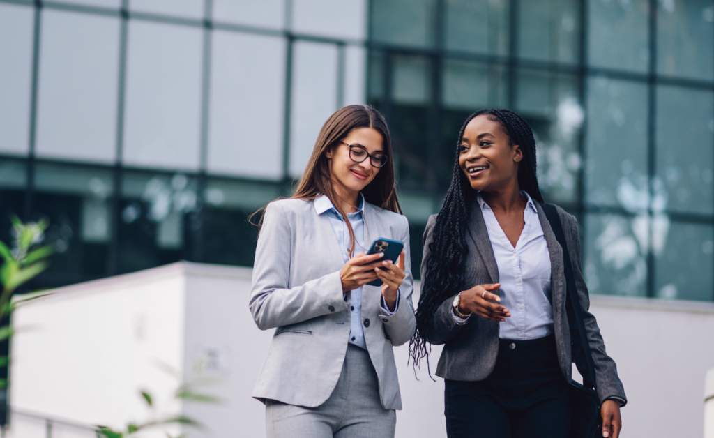Two smiling businesswomen in professional attire looking at a phone together outside an office building.