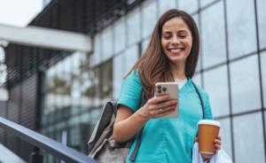 Smiling nurse in scrubs checking her phone while holding coffee, walking outside a hospital building.