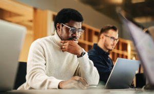 Business professional focused on his laptop screen in an office, with a colleague working nearby.