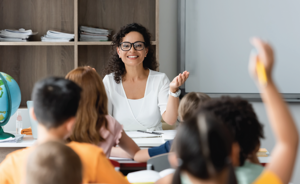 Smiling teacher gesturing while calling on a student with a raised hand in a classroom.