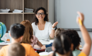 Smiling teacher gesturing while calling on a student with a raised hand in a classroom.