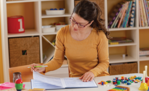 Teacher in a colorful classroom reviewing a lesson planner surrounded by learning tools and toys.
