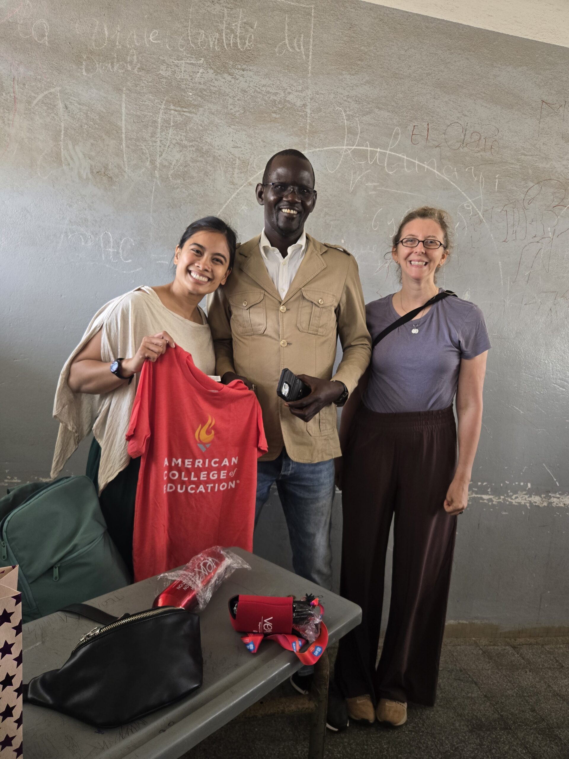Three people smiling together indoors, one holding up a red American College of Education T-shirt.