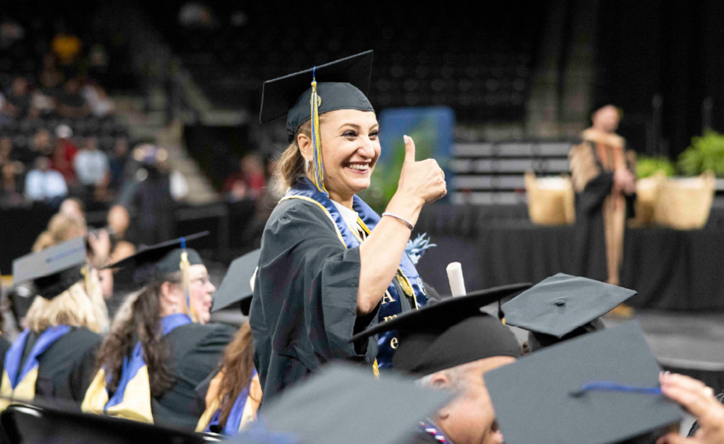 Smiling graduate in cap and gown giving a thumbs up during a college graduation ceremony.
