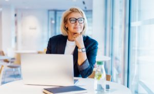 Businesswoman in a blazer sitting thoughtfully at a laptop in a bright office setting.