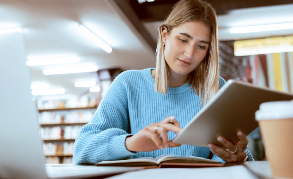 Student in a library using a tablet with an open book and laptop nearby.