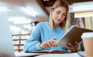 Student in a library using a tablet with an open book and laptop nearby.