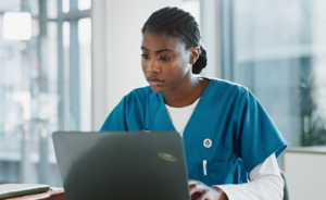 Nurse in scrubs focused on her laptop while working at a desk in a bright office.