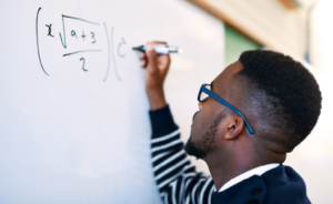 Student writing an algebraic equation on a whiteboard while wearing glasses.