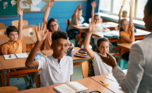 Elementary students raise their hands to answer a teacher's question in a classroom.