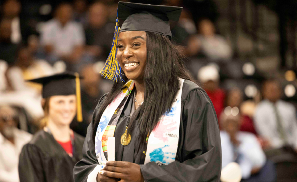 Smiling graduate in cap and gown holding a diploma at a graduation ceremony.