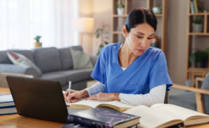 Nurse in scrubs studying textbooks and taking notes at a laptop in a living room.