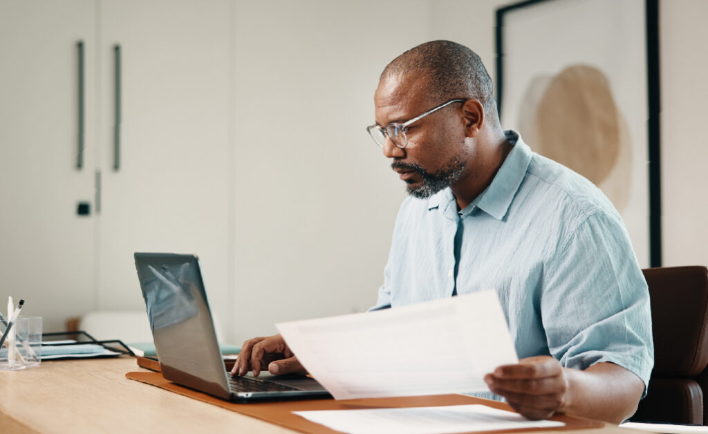 Man in an office reviewing a printed document while typing on a laptop.