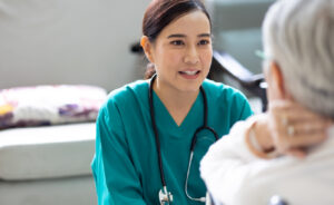 Nurse in scrubs with a stethoscope speaks with an older patient during a care visit.